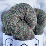 Three skeins of green yarn with speckles on white labels with a logo, against a white background.